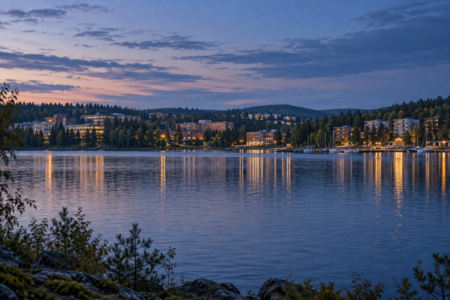 Fiktives finnisches Stadtufer am See in der Abenddämmerung mit Häusern, Bäumen und Lichtern